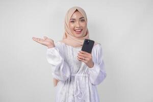 Smiling Asian woman wearing hijab and white dress using smartphone while pointing to the copy space on her right side, standing over isolated white background. Ramadan and Eid Mubarak concept photo