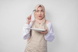 Confused young Asian Muslim woman in hijab and cream apron holding spoon and empty plate with copy space over it, thinking hard what food menu to eat for iftar. Ramadan concept photo