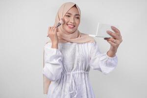 Beautiful young Asian Muslim woman wearing white dress and hijab applying make up using brush. Fashion and cosmetics beauty concept photo