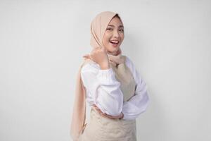 Excited young Asian Muslim woman in hijab and cream apron pointing to the empty copy space on the left and right side. Ramadan advertisement concept photo