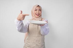 Portrait of a cheerful young Asian Muslim woman in a veil hijab and cream apron pointing to an empty plate with copy space over it, presenting food menu photo