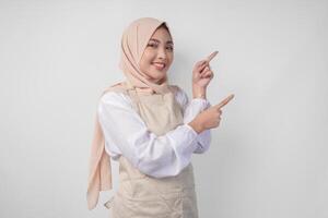 Excited young Asian Muslim woman in hijab and cream apron pointing to the empty copy space on the left and right side. Ramadan advertisement concept photo