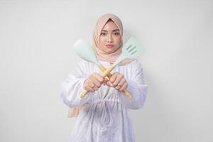 Displeased Asian woman in veil hijab doing a refusal or rejection sign, saying no, asking to stop gesture using spatula and cooking utensils photo