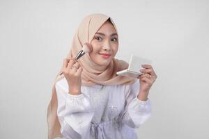 Beautiful young Asian Muslim woman wearing white dress and hijab applying make up using brush. Fashion and cosmetics beauty concept photo