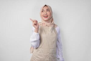 Excited young Asian Muslim woman in hijab and cream apron pointing to the empty copy space on the left and right side. Ramadan advertisement concept photo