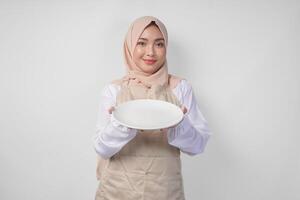 hermosa joven asiático musulmán mujer en hijab y crema delantal presentación un vacío plato con Copiar espacio terminado él, demostración el comida menú para iftar foto