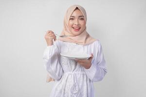 Hungry young Asian Muslim woman in hijab holding spoon and eating from an empty plate with copy space over. Ramadan concept photo