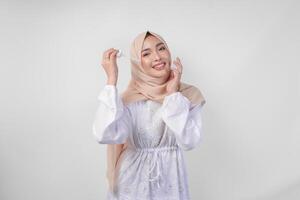 Excited young Asian Muslim woman wearing white dress and hijab testing or applying skin care serum on face. Facial and beauty concept photo