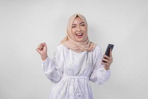 Young Asian Muslim woman with a happy successful expression wearing white dress and hijab holding smartphone, isolated by white background. Ramadan and Eid Mubarak concept photo