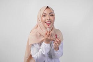 hermosa joven asiático musulmán mujer vistiendo blanco vestir y hijab poniendo en maquillaje aplicando lápiz labial. Moda y productos cosméticos belleza concepto foto