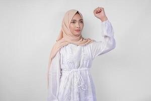 joven asiático musulmán mujer vistiendo blanco vestir y hijab con grave expresión levantamiento un apretado puño gesto terminado aislado blanco antecedentes foto