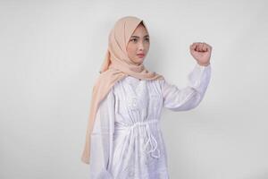 Young Asian Muslim woman wearing white dress and hijab with serious expression raising a clenched fist gesture over isolated white background photo