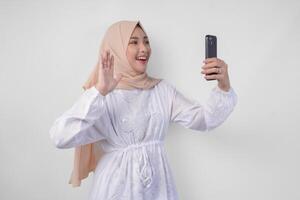 Portrait of Asian Muslim woman wearing white dress and hijab holding her smartphone to do a call to family or friend, isolated by white background. Ramadhan and Eid Mubarak concept photo