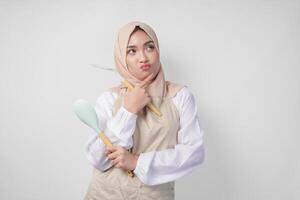 Thoughtful young Asian Muslim woman in hijab and cream apron holding spatula and kitchen cooking utensils, thinking hard what food menu to cook photo