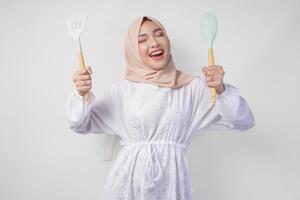 retrato de un contento asiático musulmán mujer en velo hijab participación espátula y cocina Cocinando utensilios mientras sonriente alegremente Ramadán concepto foto