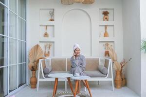 Joyful smiling Asian woman in grey bathrobe and white towel on head sitting in a couch and talking on smart phone photo