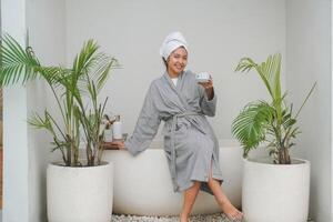 Happy attractive Asian woman in grey bathrobe posing at the bathtub, smiling cheerfully while drinking tea. Holiday leisure concept. photo