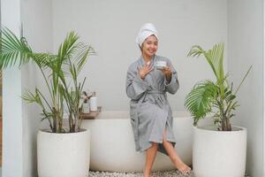 Happy attractive Asian woman in grey bathrobe posing at the bathtub, smiling cheerfully while drinking tea. Holiday leisure concept. photo