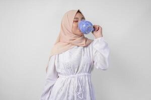 sediento joven asiático musulmán mujer en blanco vestir y crema velo hijab Bebiendo Fresco mineral agua desde un azul taza después ayuno. Ramadán concepto foto