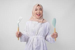 Portrait of a happy Asian Muslim woman in veil hijab holding spatula and kitchen cooking utensils while smiling cheerfully. Ramadan concept photo