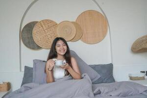 Happy attractive Asian woman in white top posing at the bedroom after waking up, smiling cheerfully while holding cup of tea. Holiday leisure concept. photo