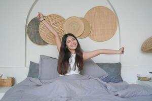 Beautiful Asian woman waking up from sleep lying on bed in bedroom, stretching hands and smiling happily photo