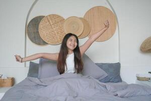 Beautiful Asian woman waking up from sleep lying on bed in bedroom, stretching hands and smiling happily photo