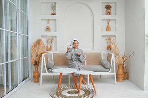 alegre asiático mujer en gris bata de baño posando a el terraza, sonriente a cámara mientras participación té taza y platillo. fiesta ocio concepto. foto