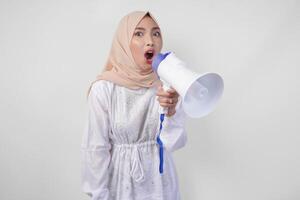 Overjoyed Asian woman wearing hijab and white dress shouting to the megaphone making announcement, standing over isolated white background photo
