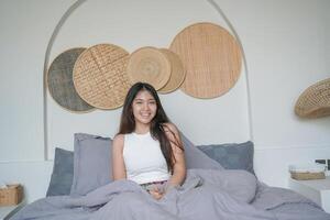 Happy beautiful Asian woman, waking up in bed and enjoys morning, looking at the camera with sleepy smile on her face photo