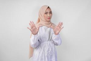 joven asiático musulmán mujer vistiendo blanco vestir y hijab demostración su palma a hacer un detener gesto con grave expresión foto