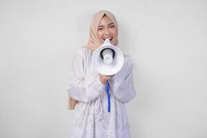 Overjoyed Asian woman wearing hijab and white dress shouting to the megaphone making announcement, standing over isolated white background photo