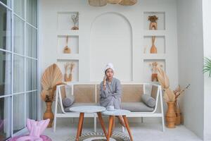 Thoughtful Asian girl in grey bathrobe sitting on a couch while thinking and having conversation on a phone call photo