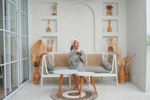 Happy attractive Asian woman in grey bathrobe posing at the terrace, smiling at camera while drinking tea. Holiday leisure concept. photo