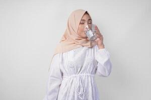 Thirsty young Asian Muslim woman in white dress and cream veil hijab drinking fresh mineral water after fasting. Ramadan concept photo