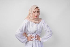 Young Asian Muslim woman in white dress and cream veil hijab holding her stomach feeling hungry during fasting. Ramadan concept photo