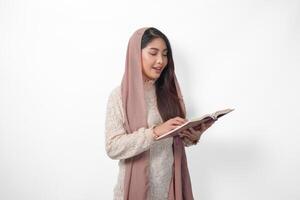 Serious Asian Muslim woman wearing veil hijab praying and reading Al Quran, standing over isolated white background. Ramadan and Eid Mubarak concept photo