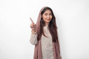 Portrait of attractive Asian Muslim woman wearing veil hijab giving number one to ten by hand gesture, standing over isolated white background photo