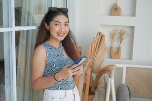 Portrait of a cheerful young Asian woman standing at terrace, smiling and holding smartphone while wearing casual outfit on holiday photo