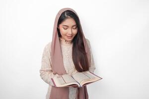Serious Asian Muslim woman wearing veil hijab praying and reading Al Quran, standing over isolated white background. Ramadan and Eid Mubarak concept photo