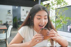 hermosa asiático joven mujer comiendo delicioso Pastelería llamado cromboloni a un cafetería. el dama picaduras pedazo de chocolate relleno cromboloni, mirando contento a el café foto