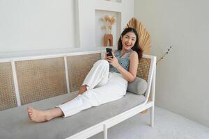 Portrait of happy Asian woman smiling and holding her smartphone while laying down on a sofa in villa for summer holiday photo