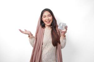 Smiling Asian Muslim woman is holding a glass of water in fasting month over isolated white background. Ramadan and Eid Mubarak concept photo