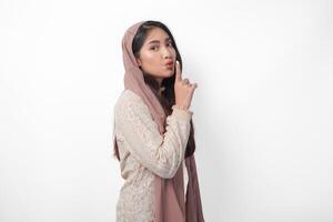 joven asiático musulmán mujer vistiendo Pañuelo velo hijab mirando a cámara mientras poniendo un dedo en frente de labios gesticulando permanecer silencio o permanecer tranquilo, aislado por blanco antecedentes foto