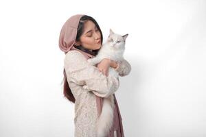 atractivo joven asiático musulmán mujer en velo hijab sonriente mientras posando y abrazando un blanco muñeca de trapo gato mascota foto