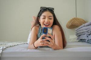 Smiling Asian woman laying down on bed with sunglasses on top while using mobile phone, exploring social media in the morning photo