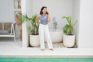 contento asiático mujer en pie por el piscina mientras vocación y hablando en un móvil teléfono con un alegre sonrisa en el cara foto