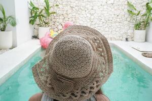 Back portrait of young Asian woman wearing white top with light cream colored hat sitting by swimming pool in a villa, relaxing and enjoying view for summer vacation photo
