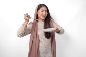 Portrait of a hungry Asian Muslim woman holding cutlery to eat or taste delicious dishes on the copy space over the plate, isolated white background photo