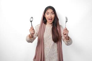 Shocked Asian Muslim woman in headscarf veil hijab holding cutlery on both hands and feeling surprised, standing over isolated white background photo